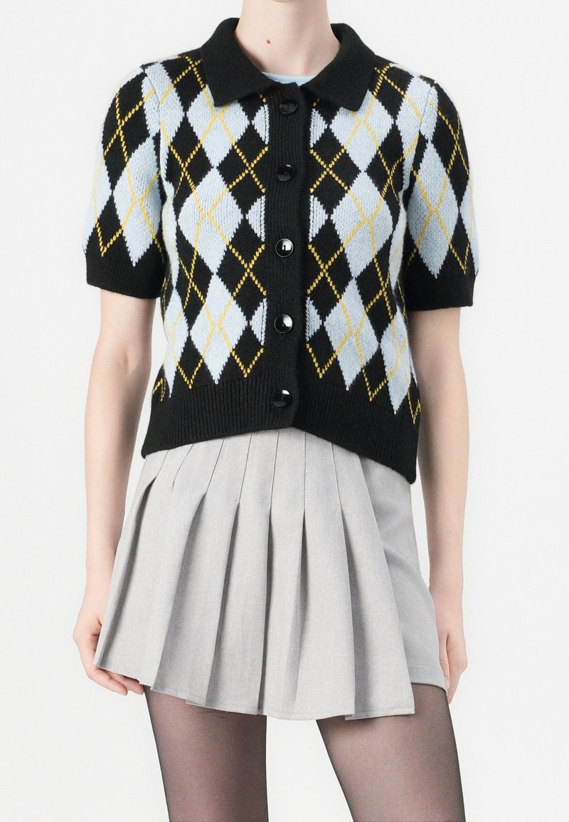 Cardigan noir à manches courtes avec boutons, motif losanges bleu et jaune, porté avec une jupe plissée gris clair et des collants noirs.