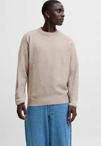 Beige stickad tröja med rund halsringning och ribbade ärmslut, kombinerad med lösa blå jeansbyxor. Mjuk textur, avslappnad passform.