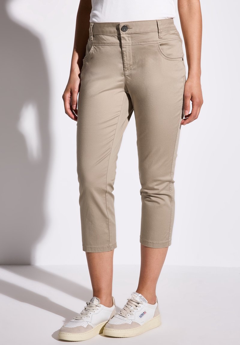 Personne portant un pantalon court beige et des baskets blanches, debout devant un fond clair uni.