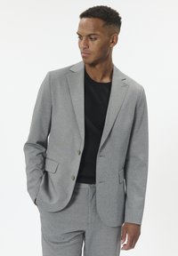 Vyras, vilkintis šviesiai pilką kostiumą ir juodą marškinių, žiūri į kairę, viena ranka kišenėje, prieš baltą fono.