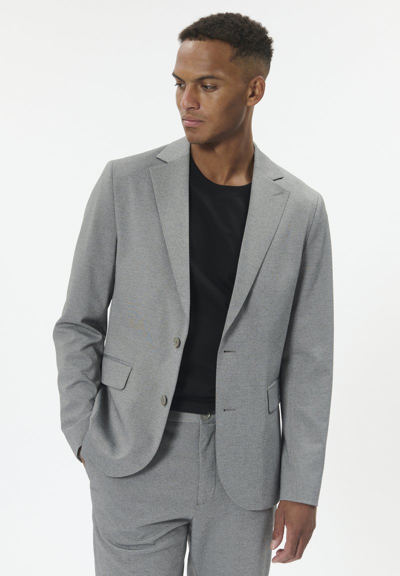 Vyras, vilkintis šviesiai pilką kostiumą ir juodą marškinių, žiūri į kairę, viena ranka kišenėje, prieš baltą fono.