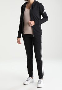 Haut à capuche noir zippé avec logo blanc sur la manche, associé à un jogging noir avec des rayures blanches sur le côté et un débardeur beige ajusté.