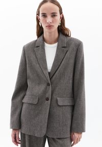 Ruskoneinen herringbone-tyylinen bleiseri, jossa on istuva muotoilu. Bleiserissä on kaksi etutaskua, kaksi ruskeaa nappia ja lovetut liepeet. Kankaan pinta on teksturoitu.