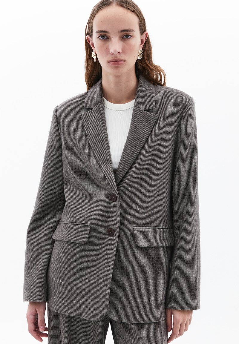 Ruskoneinen herringbone-tyylinen bleiseri, jossa on istuva muotoilu. Bleiserissä on kaksi etutaskua, kaksi ruskeaa nappia ja lovetut liepeet. Kankaan pinta on teksturoitu.