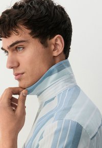 Ovratna majica iz svetloplavog platna s vertikalnimi črtami različnih širín. Ovratnik je zavihnjen navzgor; platno ima gladko teksturo.