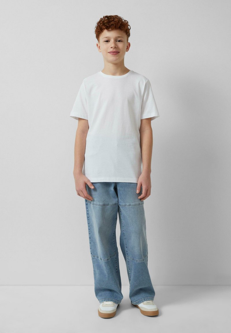 Witte t-shirt met korte mouwen, gecombineerd met lichtblauwe wijde jeans met zichtbare stiksels, en witte sneakers met beige zolen.