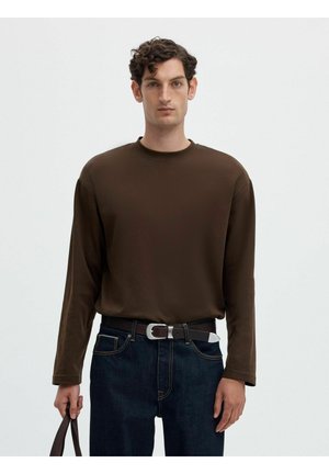 Homme aux cheveux bouclés portant une chemise marron à manches longues, un jean foncé avec ceinture, tenant un sac marron à ses côtés.