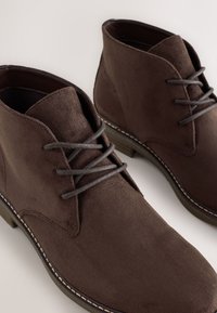 Hnedé semišové členkové topánky s okrúhlou špičkou, sivými šnúrkami a kontrastným šitým lemom pozdĺž podrážky. Textúrovaný povrch a uhladený dizajn.