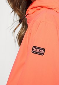 Helle orange wasserdichte Jacke mit verstellbarem Kapuze, ausgestattet mit einem schwarzen Stoff-Logopatch am Ärmel. Glatte Textur und modernes Design.