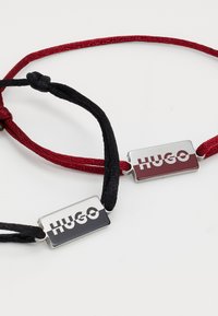Deux bracelets en cordon tressé, un noir et un rouge, chacun avec une plaque en métal mat gravée "HUGO."