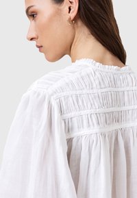 Blusa bianca con dettagli arricciati e scollatura a volant. Il tessuto è leggero e con texture, che mette in evidenza sottili pieghe verticali.