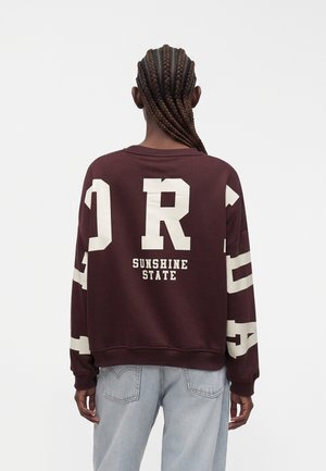 Personne aux cheveux tressés portant un sweat-shirt bordeaux avec de grandes lettres blanches et le texte « Sunshine State » dans le dos, associé à un jean bleu clair.