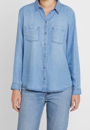 Femme portant une chemise en denim bleu clair avec boutons sur le devant et poches poitrine, assortie à un jean bleu clair, debout devant un fond uni.