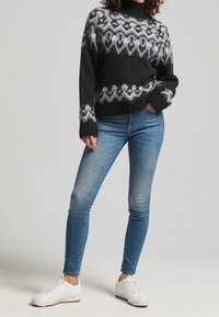 Vrouw in een zwart-witte gebreide trui met patroon, blauwe skinny jeans en witte sneakers, met handen in de zakken tegen een neutrale achtergrond.