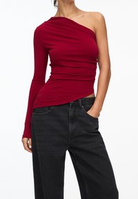 Femme portant un haut long à une épaule bordeaux et un jean noir ample, avec une main dans la poche, sur fond blanc.