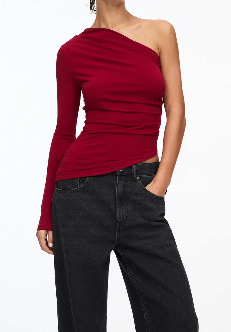 Femme portant un haut long à une épaule bordeaux et un jean noir ample, avec une main dans la poche, sur fond blanc.