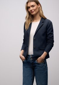 Jeune femme aux longs cheveux blonds, debout les mains dans les poches, portant un blazer bleu marine texturé, une chemise blanche et un jean bleu, sur un fond uni.