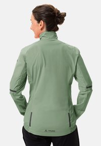 Giacca impermeabile verde con colletto alto, maniche lunghe, polsini elasticizzati e dettagli riflettenti sulle spalle e sulle tasche. Logo visibile.