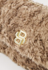Close-up van een beige, donzig textiel met een glanzend gouden dubbele-B logo en een gouden kettingdetail.