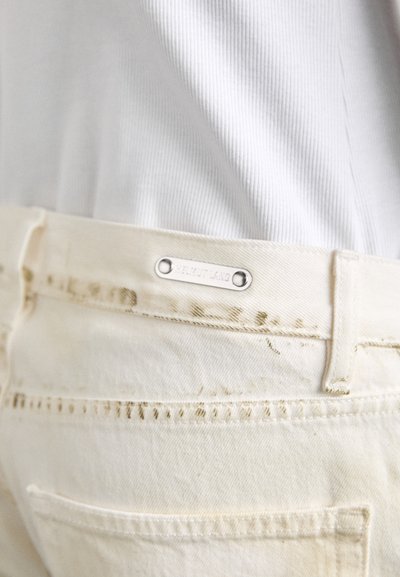 Pantalon en denim blanc avec des détails de couture contrastants, comportant une étiquette métallique embossée "HELMUT LANG" sur la taille.