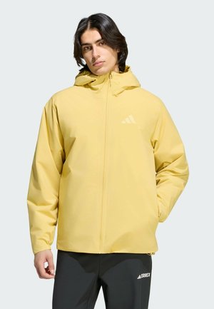 Junger Mann mit mittellangen dunklen Haaren, trägt eine gelbe Kapuzenjacke und schwarze Hose, Hände in den Taschen, stehend vor hellem Hintergrund.