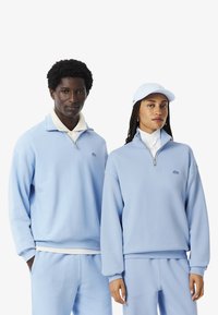 Sudaderas con cremallera de color azul claro, hechas de tela suave, con puños y cuellos acanalados, llevadas con pantalones a juego y una gorra.