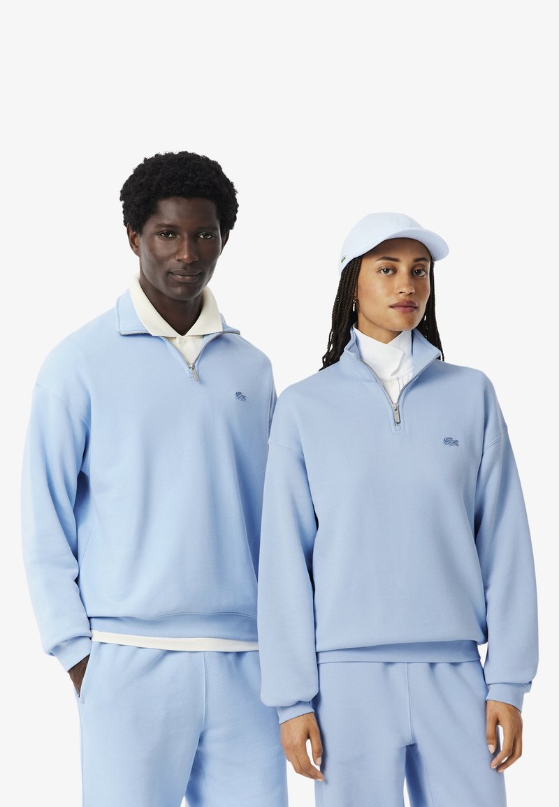 Sudaderas con cremallera de color azul claro, hechas de tela suave, con puños y cuellos acanalados, llevadas con pantalones a juego y una gorra.