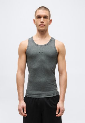 Homme aux cheveux courts portant un débardeur gris ajusté Nike et un short noir, debout devant un fond clair uni.