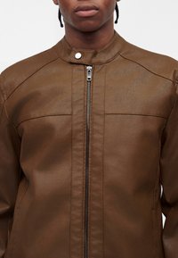 Personne portant une veste en cuir marron avec une fermeture éclair à l'avant et un col à bouton-pression, montrée du cou à la taille.
