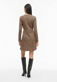 Robe marron côtelée à manches longues, taille cintrée, jupe évasée et encolure ronde, associée à des bottes noires montantes jusqu'aux genoux.