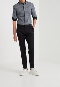 Camicia button-up blu con un piccolo motivo geometrico bianco, abbinata a pantaloni slim-fit neri e sneakers bianche.
