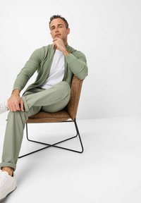 Light green zip-up cardigan over a white shirt, paired with light green pants and white sneakers. Seated on a brown accent chair.