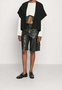 Czarny dzianinowy krótki kardigan na zielonej prążkowanej bluzce, zestawiony z czarnymi skórzanymi szortami i błyszczącymi czarnymi loafersami z srebrnymi akcentami.