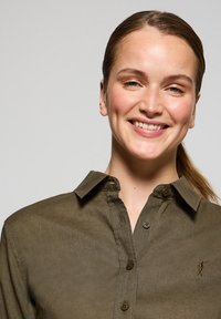 Olivgrön skjorta med krage och knappar, med mörka knappar och en liten broderad logotyp på vänster sida. Slät textur.