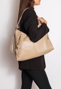 Beige leren handtas met een gestructureerde afwerking, voorzien van gouden hardware en een zijrits. Versierd met een decoratieve hondvormige bedel.