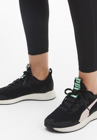 Zapatos deportivos negros con parte superior de malla, entresuela blanca, acentos en verde claro y un panel lateral rosa. Presentan cierre de cordones y superficie texturizada.