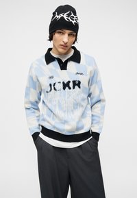 Νεαρός που φοράει μαύρο σκουφάκι και ανοιχτό μπλε και λευκή καρό μπλούζα με μαύρο γιακά και γράμματα, στέκεται με τα χέρια στις τσέπες.