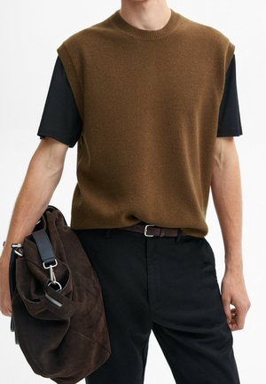 Mężczyzna w brązowym bezrękawniku na czarnym t-shircie, czarne spodnie, trzyma ciemnobrązową zamszową torbę z paskiem.