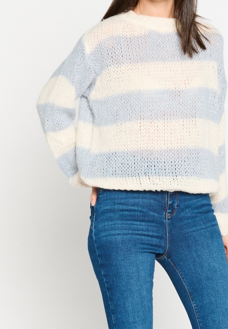 Femme portant un pull en maille ample avec de larges rayures horizontales crème et bleu clair, associé à un jean bleu ajusté, main dans la poche.