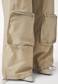 Beige Cargo-Hosen mit großen Reißverschlusstaschen, weiter Passform und glatter Textur, mit verstellbaren Bündchen und spitzen beigen Schuhen darunter.