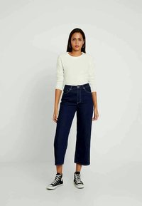 Jeans de cintura alta azul escuro com um corte relaxado, bolsos frontais e costura branca, combinados com um suéter leve de manga longa e ténis pretos.