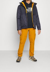 Muž oblečený v černožluté vrstvené bundě, černé mikině The North Face, hořčičkových kalhotách a béžovočerných teniskách, stoj ící na bílém pozadí.