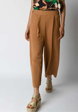 Tan, pleated, ankle-length trousers made of lightweight fabric, paired with gold-toned platform sandals, featuring two straps over the foot.