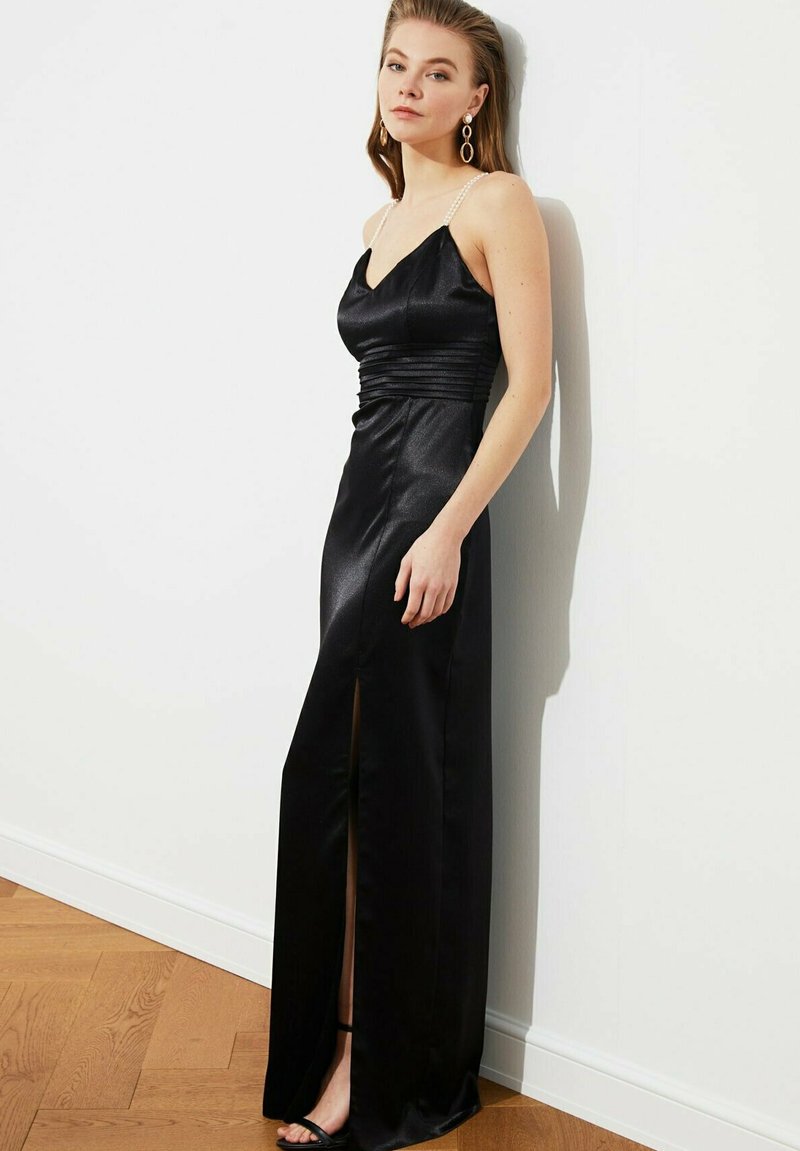 Czarna satynowa sukienka wieczorowa z głębokim dekoltem w kształcie V, marszczoną talią i rozcięciem do uda. Posiada ramiączka na cienkich, perlitych paskach oraz gładką strukturę.