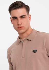 Světle hnědá polo triko s límečkem a krátkými rukávy. Na hrudi má černou trojúhelníkovou nášivku. Hladká textura.