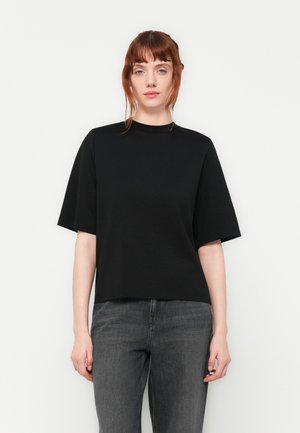 Ung kvinne med rødt hår iført en svart t-skjorte med korte ermer og grå jeans, stående mot en enkel hvit bakgrunn.