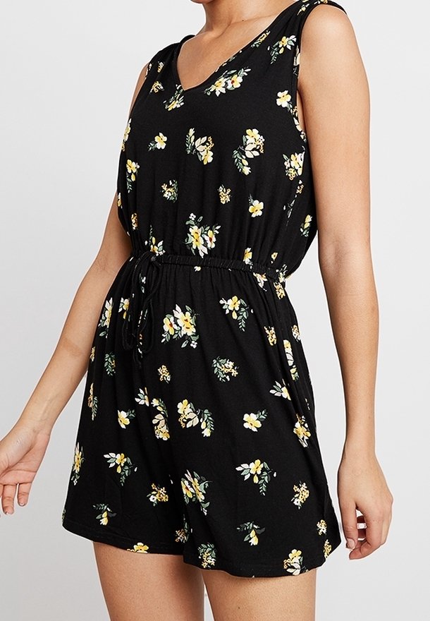 Combinaison noire sans manches avec imprimé floral jaune et blanc, taille élastique avec cordon, portée par une personne debout sur un fond uni.