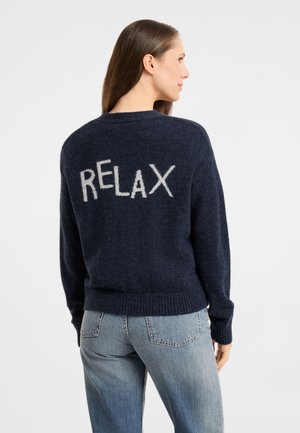 Frau mit langen braunen Haaren, die einen dunkelblauen Pullover mit dem Wort "RELAX" auf dem Rücken und hellblaue Jeans trägt, seitlich stehend.
