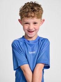 Jeune garçon souriant aux cheveux bouclés portant un maillot de sport bleu rayé Hummel Performance, les bras croisés devant un fond blanc.