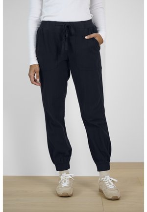 Person trägt schwarze Jogginghosen mit Kordelzug und elastischen Bündchen, weißes Langarmshirt, weiße Socken und beige Turnschuhe und steht auf einem Holzboden.